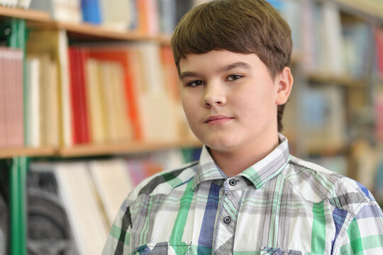 Half growth portrait of student in plaid shirt, amid shelves of books, looking at camera