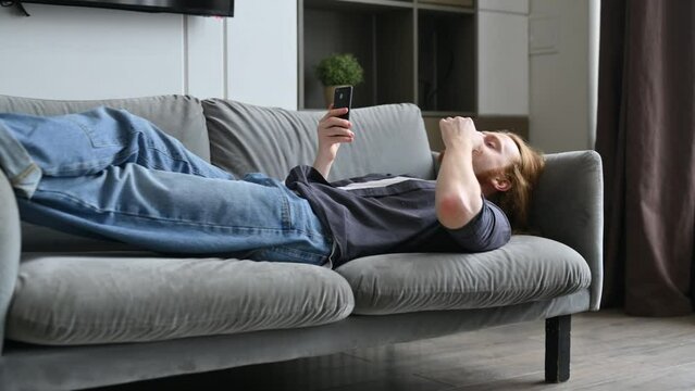 A young red-haired guy is lying on the sofa and eating corn flakes, scrolling through social networks on his phone