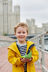 Little smiling boy in yellow jacket stands on embankment holding green paper boat in his hand