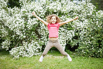 Little girl jumps outstretching arms and legs against jasmine bush in summer park