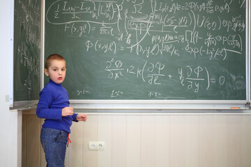 Little cute boy stands near blackboard with formulas in classroom