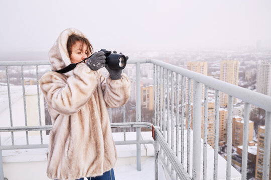 Woman in fur jacket takes pictures of city standing on building roof in winter