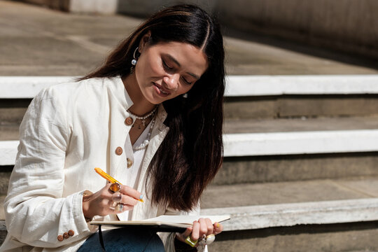 Smiling woman sitting on stairs writing on a notebook.