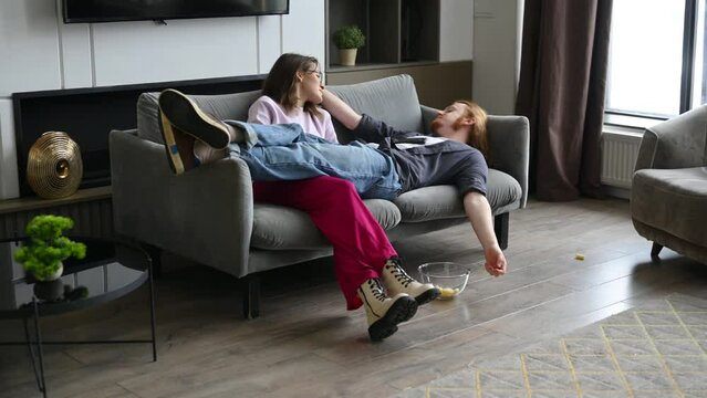 A young red-haired guy with his girlfriend is lying on the sofa and eating corn flakes and chatting