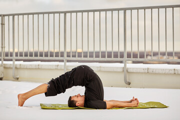 Young man does exercises on mat at snowy roof in winter