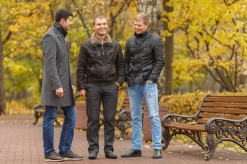 Three young men in black jackets standing near park bench, talk and smile