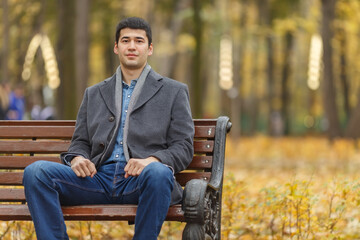 Portrait of young man in gray coat and jeans sitting on bench in alley in autumn park