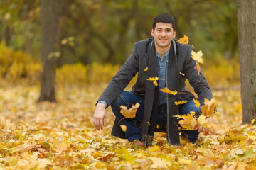 Young man in gray coat sitting on his haunches in park and pop-up Yellow maple leaves
