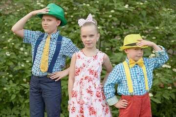 Two boys in hats and girl stand outdoor against green bushes