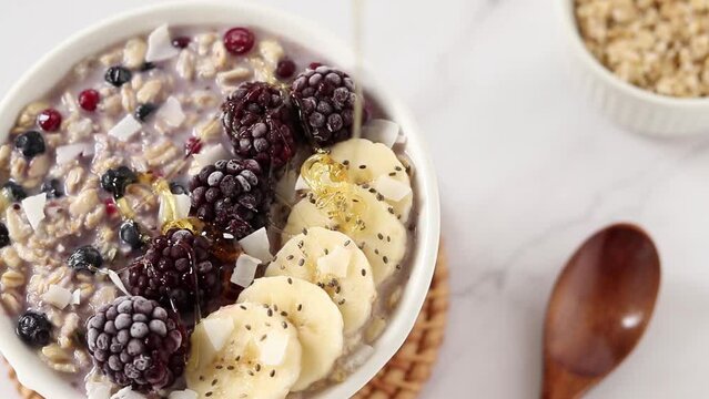 Pouring honey with wooden dipper on oatmeal breakfast bowl with berries, bananas, and chia seeds. Close-up. Healthy eating and dieting concept.
