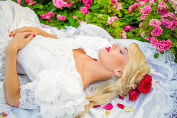 Portrait of beautiful woman in white dress with roses in her hair lying on lawn and sleeping among flowers in park