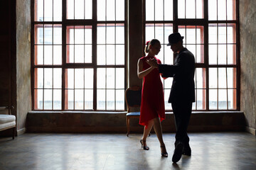 Woman in dress and man in black suit dance tango near big window in room