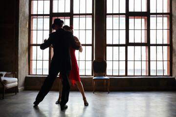 Woman in red dress and man in black suit dance tango near big window in room