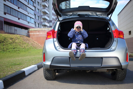 Little girl sitting in open trunk of car and looking through binoculars