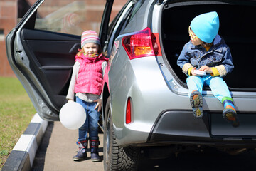 Little boy in yellow glasses sitting in open trunk of foreign car and girl with white balloon...