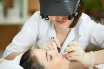 Beautician in magnifying lenses does epilation on upper lip of woman lying on couch in beauty salon.