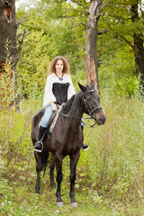 Smiling woman with curly hair sits on bay horse in the park.