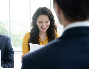 A woman in a yellow shirt is reading a piece of paper while a man in a suit watches her. The scene...