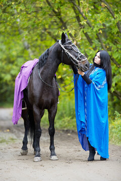 Black-haired woman in blue capote stands with horse in the park.