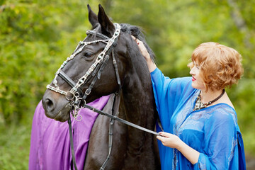 Red-haired smiling woman in blue capote with bay horse in the park.