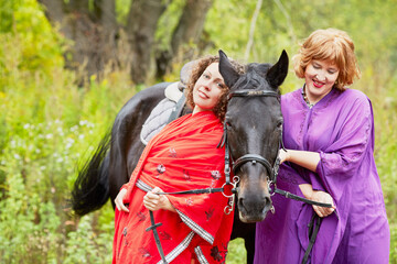 Two women in red and violet clothes with chestnut horse in park.