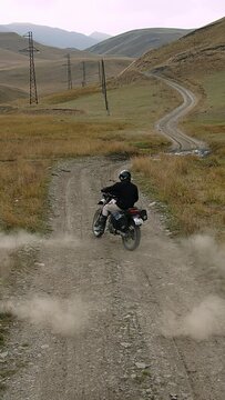 Captured in the midst of a daring maneuver, a motorcyclist clad in black with a dark helmet executes a tight circle turn on a gravel mountain path. The bike, at an angle, kicks up a thick cloud of