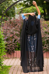 Woman with raised hands in black indian sari stands in summer garden, back view