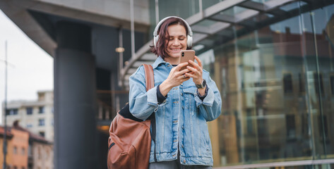 Young Caucasian happy cheerful woman in headphones listening to music using smartphone in the city