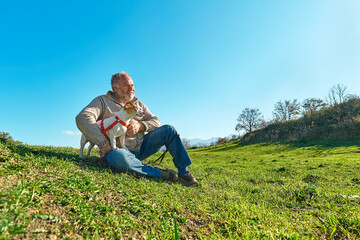 Naklejka na ściany i meble Gray haired man taking a walk with his jack russell dog in meadow in mountainous area. Mature man playing outdoors with his dog in nature.