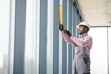 African Workman in overalls installing or adjusting plastic windows in the living room at home
