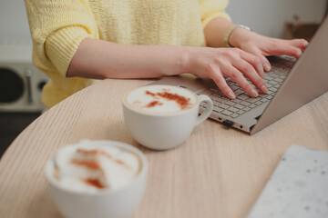 Close-up on hands typing on a laptop, beside frothy cappuccinos, coffee suggesting a productive coffee break, business and personal growth.