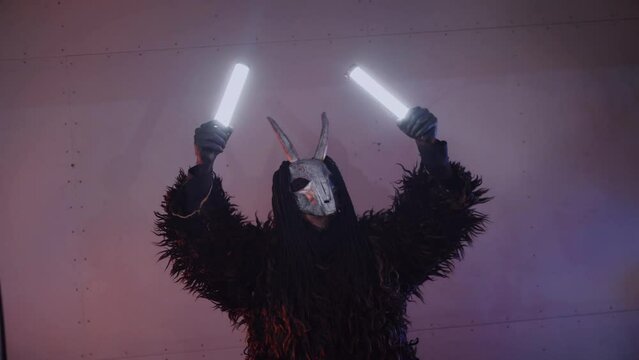 The devil monster dances with RGB sticks in his hands. A mystical man in a scary mask in a dark studio. The man is dressed in a black coat and a mask with horns on his head. Alien from space. Horror,