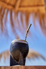 close-up of Argentine mate on a beach under a thatched umbrella and blue sky