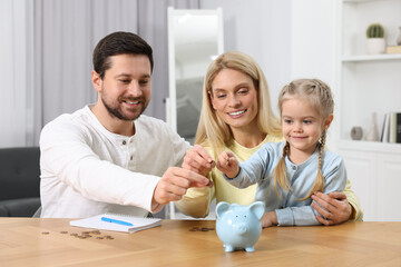 Planning budget together. Little girl with her family putting coins into piggybank at table indoors