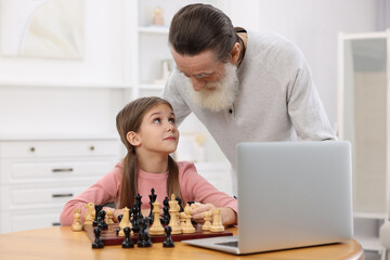 Obraz na płótnie Canvas Grandfather teaching his granddaughter to play chess following online lesson at home