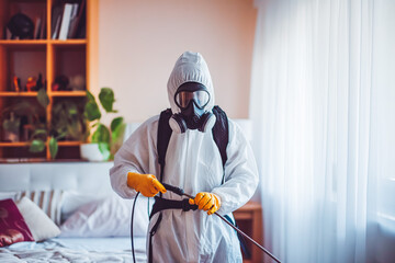 Faceless pest control worker in a protective suit sprays insect poison in bedroom. White room.