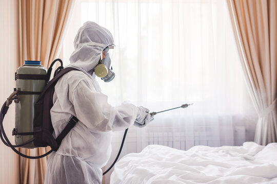 Faceless pest control worker in a protective suit sprays insect poison in bedroom. White room.