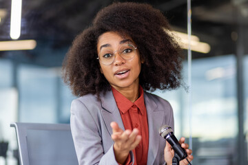 Close-up portrait of an African-American businesswoman in a suit and glasses sitting in the office at a desk, holding a microphone and talking to the camera while gesturing with her hands.