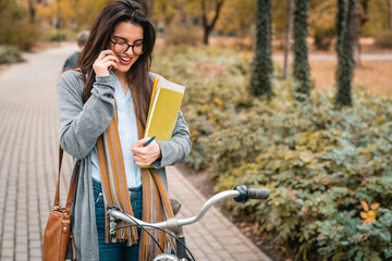 Attractive young adult business woman standing in the city park while holding notepad and having a joyful phone conversation.
