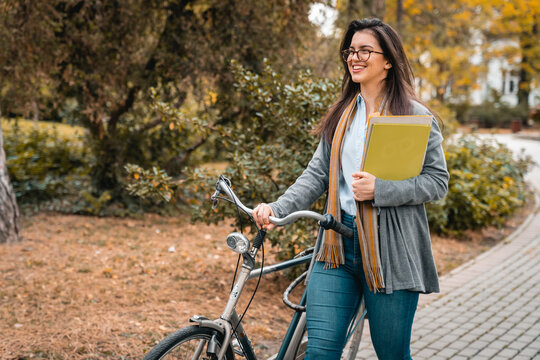 Smiling young business woman holding papers and pushing her bike while walking in public park on her way to the office.