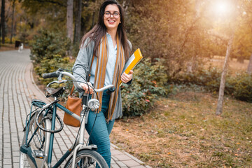Smiling young business woman holding papers and pushing her bike while walking in public park on...