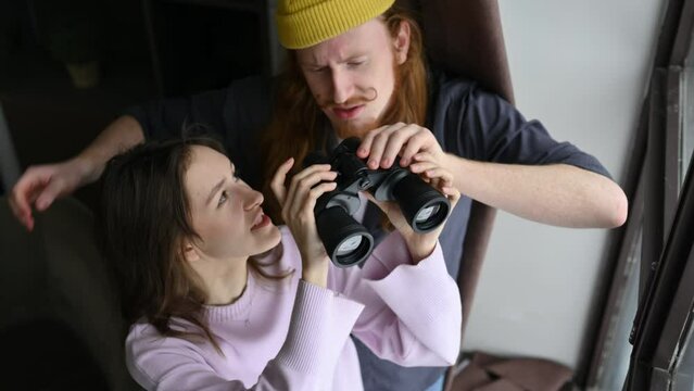 A young woman and her boyfriend are watching someone through binoculars from the window