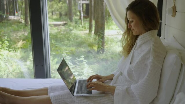 Woman using laptop sitting on bed. Young smiling cheerful girl in white bathrobe on vacation with computer on lap typing message, texting chatting online, writing blog article, working, studying at pc