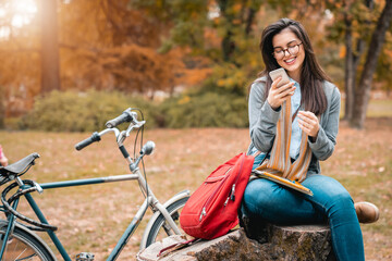 Young female student using mobile phone while studying for exam in public park during the sunny Autumn day.