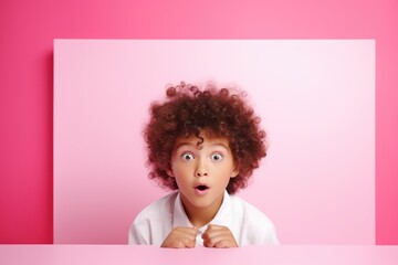 Boy with curly hair peeks over a pink surface, his eyes wide in astonishment and curiosity, embodying a moment of pure wonder and surprise - obrazy, fototapety, plakaty