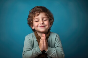 Boy with soft curls closes his eyes and clasps hands in a hopeful gesture, wearing a cozy sweater against a soothing blue background