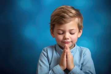 Fototapeta premium Young boy with closed eyes and clasped hands, deeply immersed in prayer or meditation, radiates peaceful contemplation, blue background