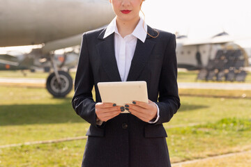 Anonymous air hostess in a tailored suit holds a tablet while standing confidently at an airfield, with a glimpse of aircraft in the background