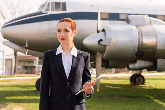 A professional air hostess stands confidently with a clipboard in hand against the backdrop of a classic propeller aircraft, symbolizing aviation expertise