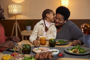 Portrait of African American little girl kissing grandma on cheek at dinner table with family copy...
